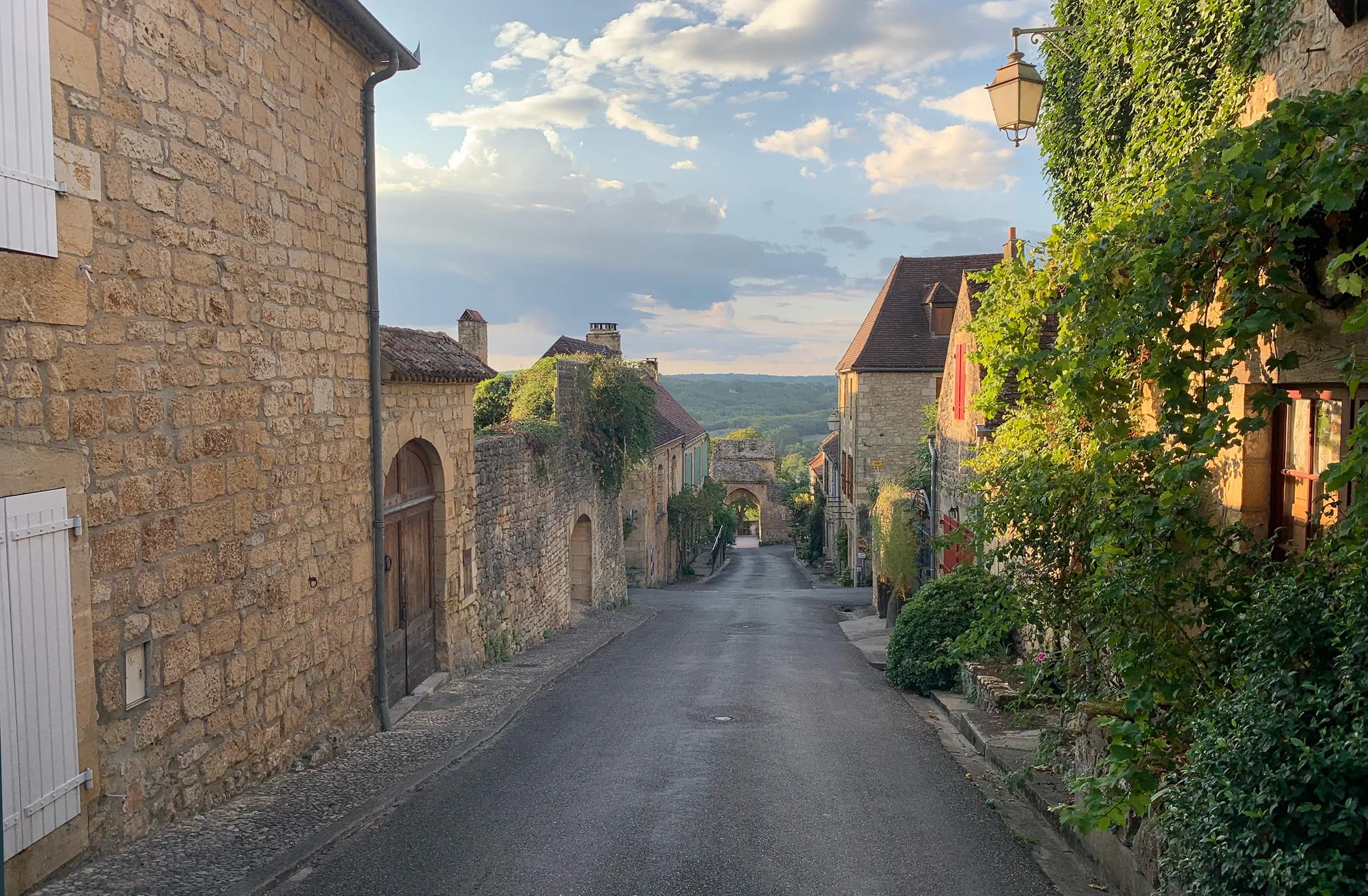A quiet lane between honey-coloured stone houses in a French hill village, ivy on the walls and wooded hills beyond